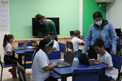 crianças estão sentadas e mexendo em computores sobre as mesas. Adultos acompanham. #paratodosverem