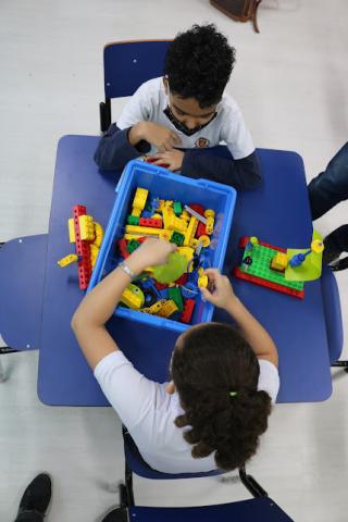 Foto do alto mostra duas crianças frente a frente com uma caixa com peças coloridas dentro. Uma delas manuseia as peças. #paratodosverem