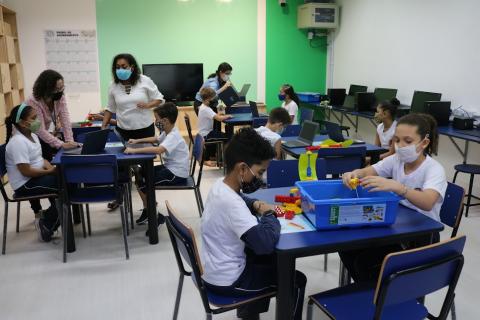 foto geral da sala com alunos sentados mexendo em computadores e caixas com peças coloridas. #paratodosverem
