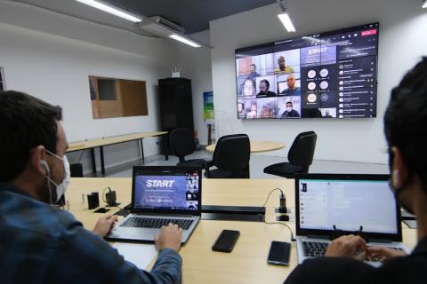 pessoas estão sentadas diante de laptops abertos e de frente para telão onde se vê várias pessoas em apresentação virtual. #paratodosverem