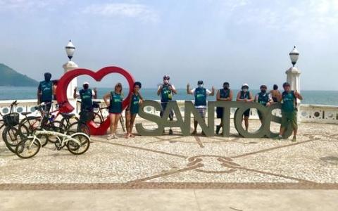 Ciclistas posam para foto no monumento Eu amo Santos na praia #paratodosverem