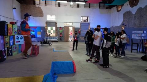 Artista fala a estudantes dentro de grande galpão decorado com pinturas em uma das paredes. #paratodosverem