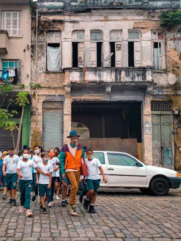 artista lidera grupod e estudantes que caminham por rua de paralelepípedo. Ao fundo, um antigo casarão. #paratodosverem