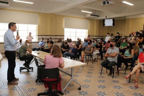 auditório com o prefeito falando ao microfone, em pé e atrás de uma mesa.  Ao lado dele há um secretário e a vice-prefeita. O auditório está cheio de pessoas sentadas. #paratodosverem