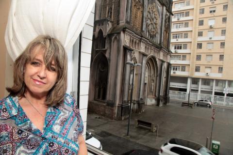 Maria de Fátima Abreu posa para foto dentro de casa, com a cortina aberta. Bem à frente se vê a fachada da Igreja de Santo Antonio do Embaré. #paratodosverem