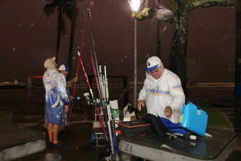 três pescadores estão em torno de mesa e cheios de apetrechos de pesca, como varas. Eles estão em área de quiosques na praia. O mar está ao fundo. É noite. #paratodosverem