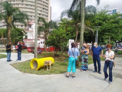 pessoas circulando nas alamedas do jardim da orla ao lado de espaço onde há peça para a distração de cães. Um cão está ao lado de uma peça. #paratodosverem