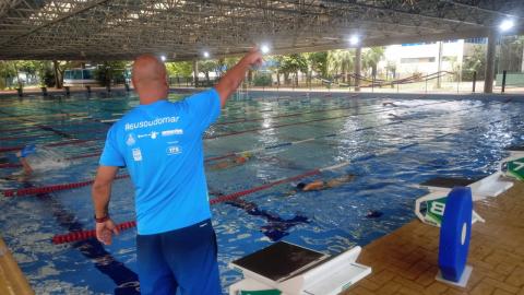 PROFESSOR, DO LADO DE FORA DA PISCINA, ESTÁ DE COSTAS PARA A IMAGEM. eLE ESTÁ COM O BRAÇO E A MÃO DIREITA ERGUIDOS, SINALIZANDO ALGO PARA QUEM ESTÁ DENTRO DA PISCINA. #PARATODOSVEREM