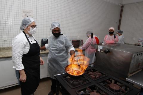 alunos e professora em torno de um fogão industrial. Destaque para a frigideira onde se prepara recheio com chama de fogo no ar