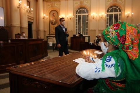 O prefeito fala em pé ao fundo da imagem. Em primeiro plano, sentada e de lado, uma menina usa roupa típica portuguesa. #paratodosverem