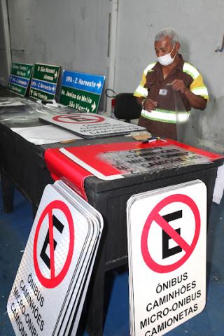 homem está atrás de mesa onde há várias placas. No chão também estão várias delas empilhadas em pé. #paratodosverem