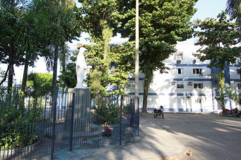 Santa está em área cercada em praça. Ao fundo, prédios de moradia. #paratodosverem