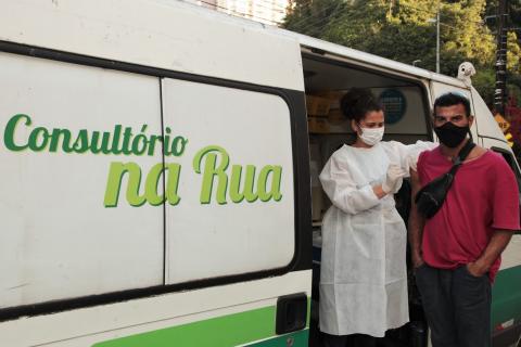 homem é vacinado por mulher. A aplicação ocorre junto a uma van onde se lê Consultório na Rua. #paratodosverem