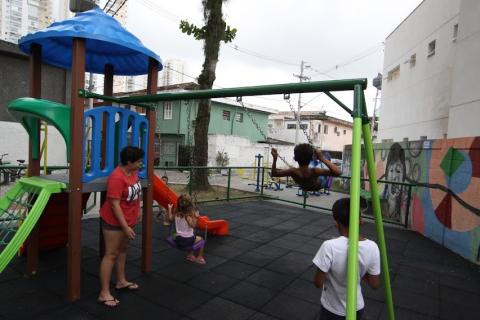 duas crianças se divertem no playground. Uma mulher acompanha. #paratodosverem
