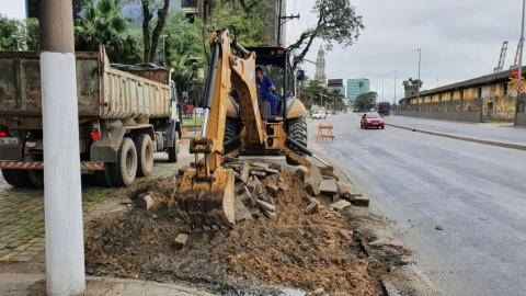 escavadeira remove parte do piso de uma praça. Há um caminhão do lado esquerdo e a pista da avenida, à direita. #paratodosverem