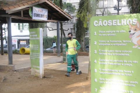 homem caminha na praça segurando uma vassoura. À direita, um banner com uma lista com Cãoselhos. #paratodosverem
