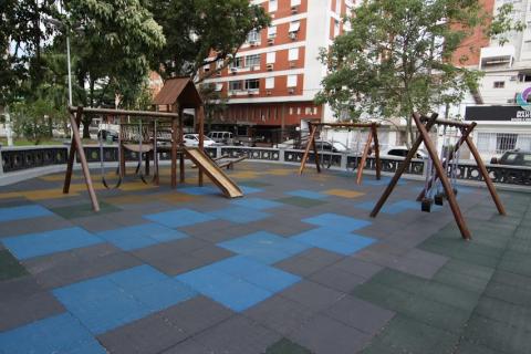 vista geral do playground com piso emborrachado e brinquedos. Há balancês e escorregador. #paratodosverem