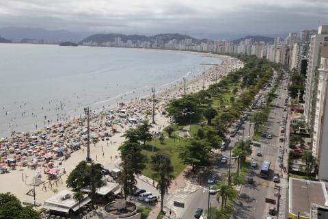 vista geral da orla cheia de gente, com a avenida, jardim, faixa de areia e mar. Imagem do alto mostra desde a ponta da praia até o José Menino.  #paratodosverem