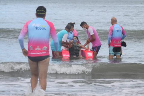 Jovem desfruta banho de mar em cadeira anfíbia com instrutores em volta. #pracegover