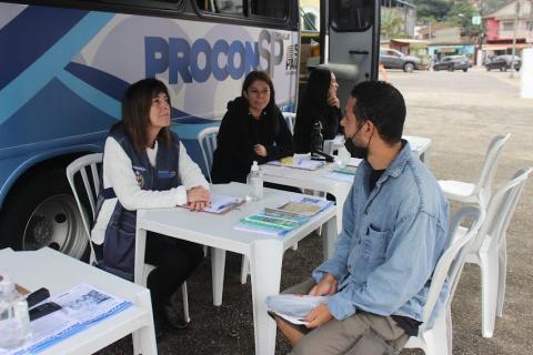 van  com lateral onde se lê procon SP. À frente, três mulheres sentadas aguardando o públicos. Um homem está sendo atendido. #paratodosverem