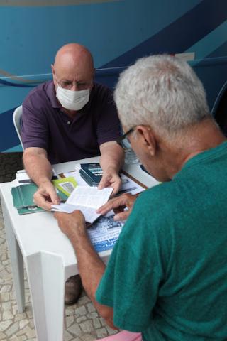 dois homens sentados à mesa, um diante do outro, seguram folheto explicativo. #paratodosverem