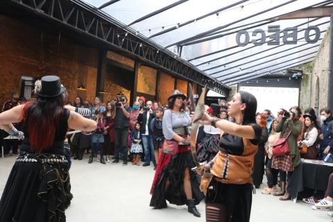 Mulheres fantasiadas dançam em apresentação no espaço de eventos. #pratodosverem