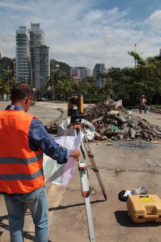 homem está atrás de aparelho de sondagem. Ele segura um papel. Ao fundo, pedras amontoadas. #paratodosverem