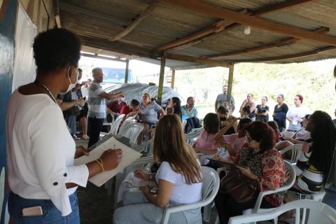 pessoas estão sentadas participando de uma reunião. Em primeiro plano e à esquerda, uma mulher em pé faz anotações em um papel sobre uma prancheta. #paratodosverem