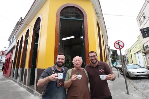 Três homens posam para foto na fachada de estabelecimento. Cada um segura uma xícara de café. #paratodosverem