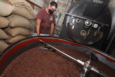 máquina de torrefação repleta de grãos. Um homem está à frente da estrutura. Ao fundo, várias sacas de café. #paratodosverem