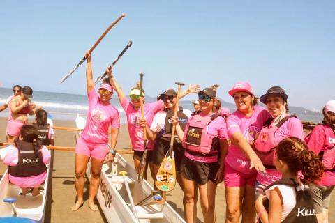 mulheres empunham remos no ar. A canoa está sobre a areia dura. O mar está ao fundo. Todas se vestem de cor-de-rosa. #paratodosverem