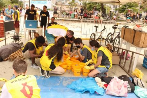 jovens usando coletes amarelos estão sentados no chão mexendo em vários baldes onde está depositado lixo. #paratodosverem