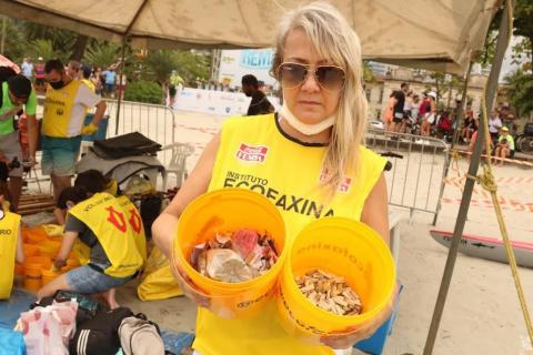 mulher com colete amarelo onde se lê Ecofaxina segura e mostra dois baldes com diversos itens inservíveis colhidos na praia. #paratodosverem