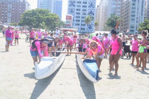 mulheres empurram canoa na areia dura. Ao fundo, o movimento da praia e os prédios da orla. #paratodosverem