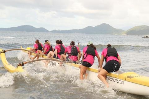 mulheres empurram canoa. Na parte da frente, algumas já estão dentro da embarcação. Outras duas, no fundo, ainda estão com as pernas esquerdas para fora. #paratodosverem