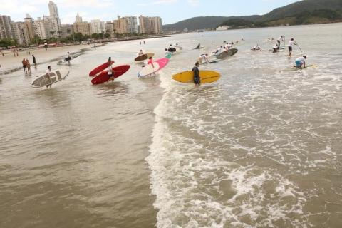 imagem de longe. Atletas ingressam no mar. Cada um segura no braço sua própria embarcação. #paratodosverem