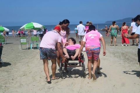 quatro pessoas empurram uma cadeira de rodas com uma mulher até a beira d'água. O mar está ao fundo. #paratodosverem