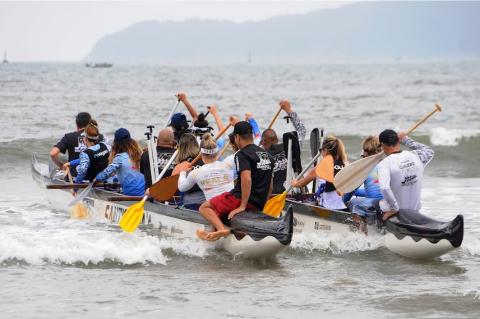 Praticantes saem para o trajeto, ainda na beira do mar, dentro da catamarã. Eles empunham remos. #paratodosverem