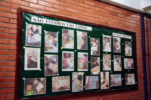 Painel afixado em parede onde se lê no topo: Não estamos tão longe. Há várias fotos de alunos estudando em casa. #paratodosverem