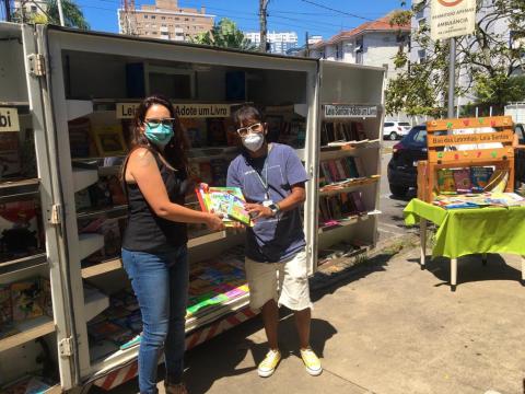 Mulher está em pé ao lado de jovem. Ambos seguram um livro. Atrás dele está o móvel onde ficam expostos os livros. #paratodosverem