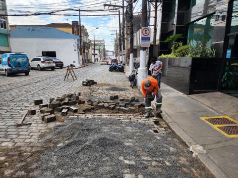 homens renivelam trecho de pista junto a meio fio. Há vários paralelepípedos soldos e pedras em torno. #paratodosverem