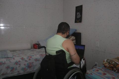 homem está sentado em cadeira de rodas, de costas para a foto. Ele está em um quarto, ao lado de uma cama. #paratodosverem