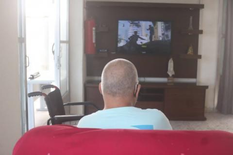 homem está de costas para a foto sentado em um sofá. Ele assiste a TV ao fundo. Ao lado esquerdo, uma cadeira de rodas. 