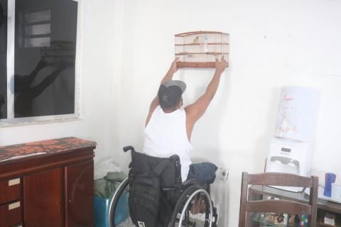 homem em cadeira de rodas está em uma sala. Ele estica os braços para o alto para pendurar uma gaiola de passarinho na parede. #paratodosverem