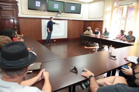 homem está em pé apontando para telão. Ele é acompanhado por várias pessoas sentadas numa mesa em formato de letra U. #paratodosverem