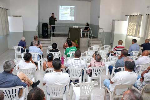 pessoas sentadas em cadeiras em um salão. Ao fundo, no alto de um tipo de palco, uma pessoa fala ao microfone diante de uma projeção. #paratodosverem