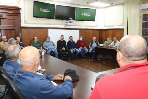 reunião com várias pessoas sentadas em torno de uma grande mesa em formato de u e outras também sentadas. #paratodosverem