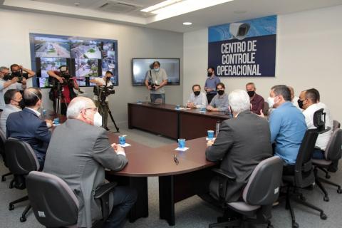 pessoas reunidas em torno de mesa. Na parede ao fundo há uma placa onde se lê: Centro de Controle Operacional. Em outra parede, monitores de tevê. #paratodosverem