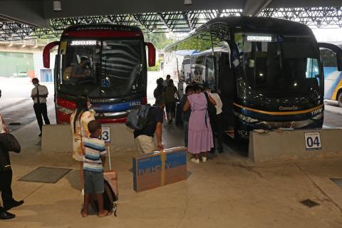 Dois ônibus parados em baias com algumas pessoas aguardando para embarcar. Há uma embalagem de televisão no chão. #paratodosverem