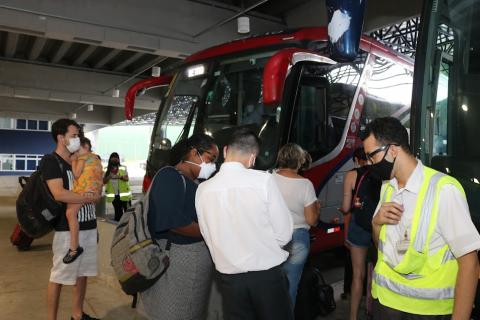 movimento de pessoas entrando em ônibus. Em primeiro plano, um homem com uma faixa reflexiva no tronco, um homem com camisa branca, de costas, uma mulher entrando no ônibus e um homem com uma criança no colo. #paratrodosverem